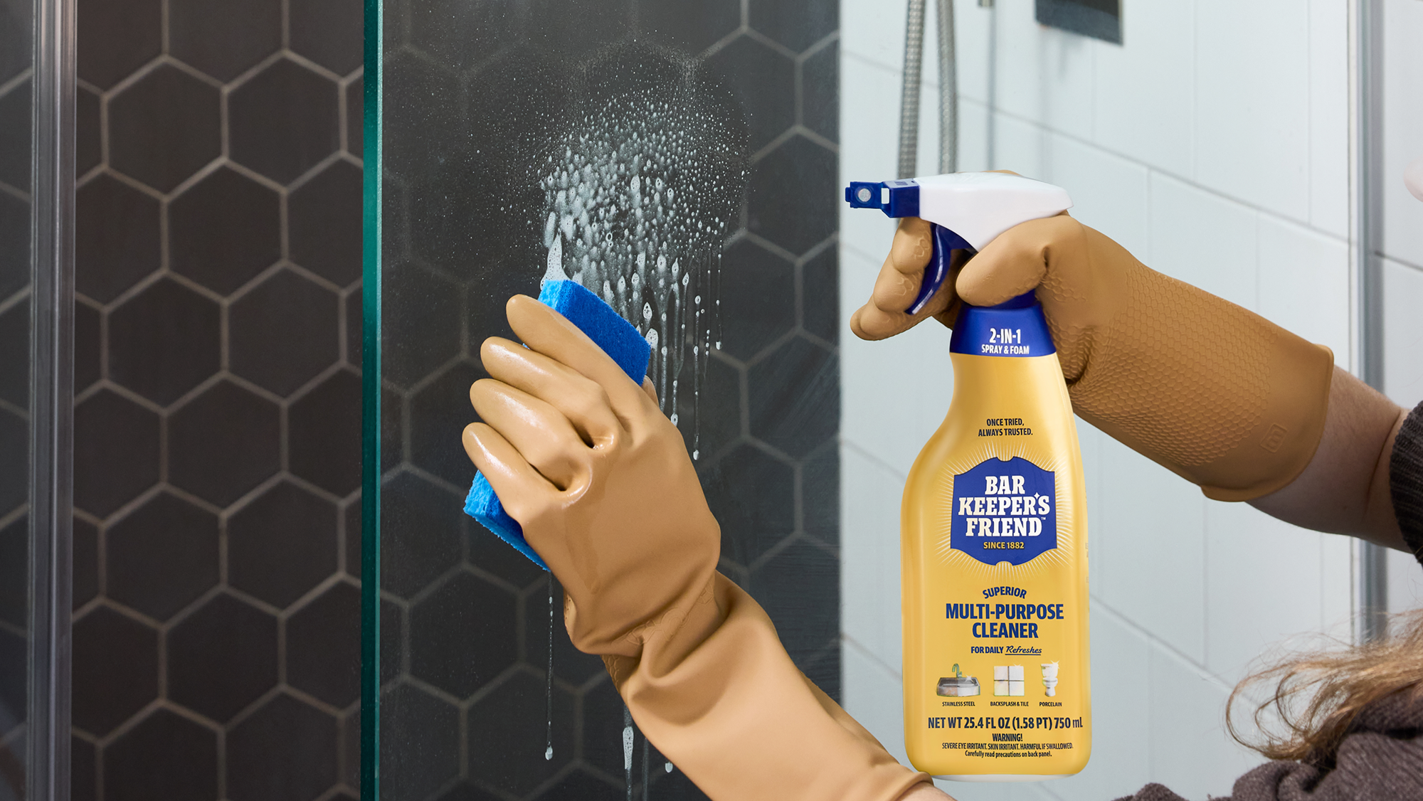 Person cleaning a glass shower door with a cleaning product in a bathroom.