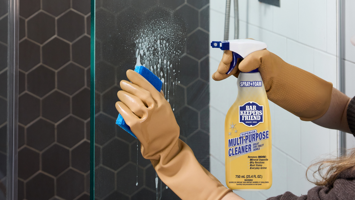 Person cleaning a glass shower door with a cleaning product in a bathroom.