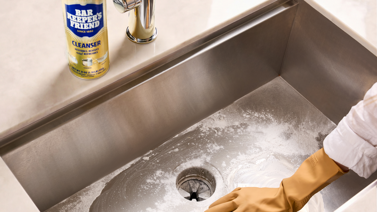 Person cleaning a kitchen sink with a sponge and wearing a glove, with a bottle of All-Purpose Friend cleaner on the counter.