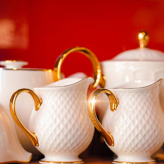 White porcelain creamers with gold handles and textured diamond pattern against a red background.