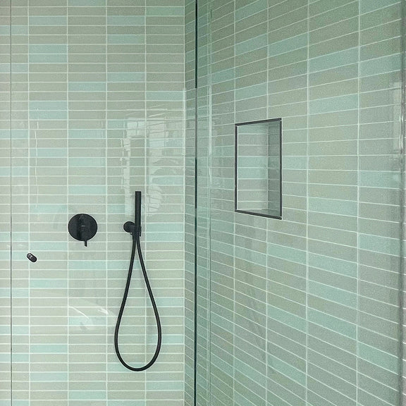 Shower area with light green rectangular tiles, black showerhead, handle, and built-in wall shelf.