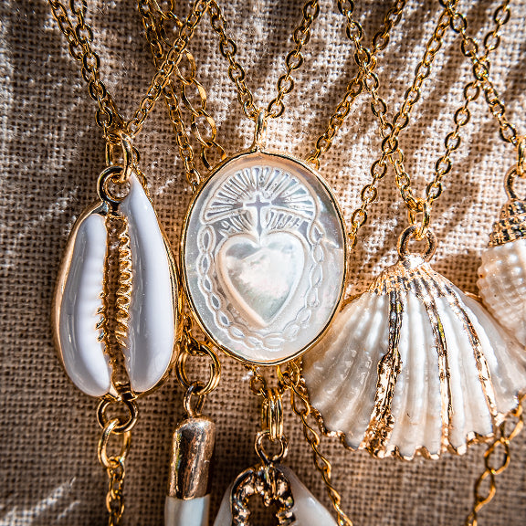 Gold chain necklaces with white shell and heart-shaped pendants on a textured fabric background.