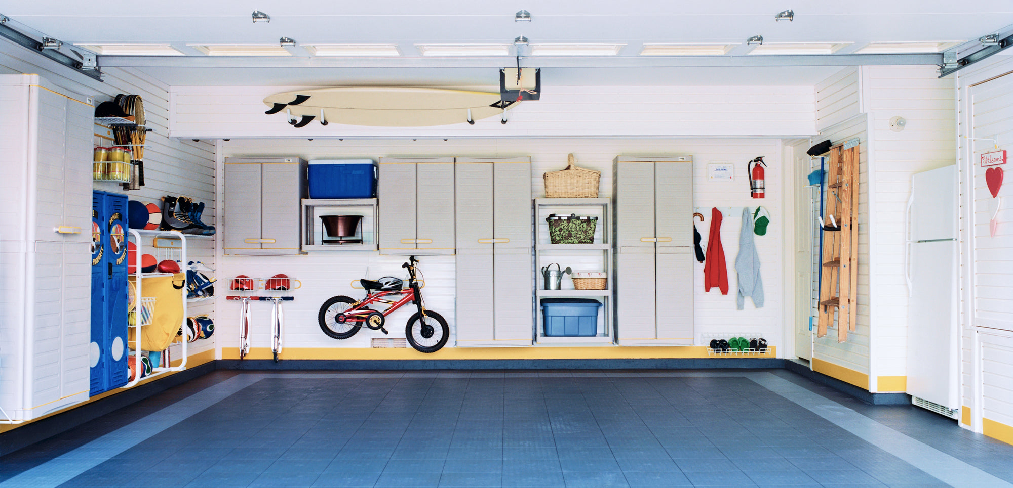 Organized garage with cabinets, shelves, a bicycle, surfboard, and hanging tools on white walls.
