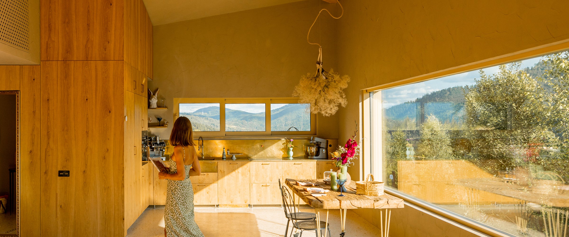 Woman standing in a modern kitchen with large windows overlooking greenery