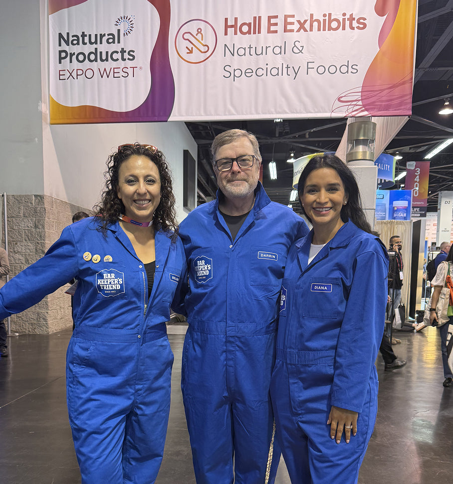 Three people in blue BKF uniforms standing in front of a 'Natural Products Expo West' banner.